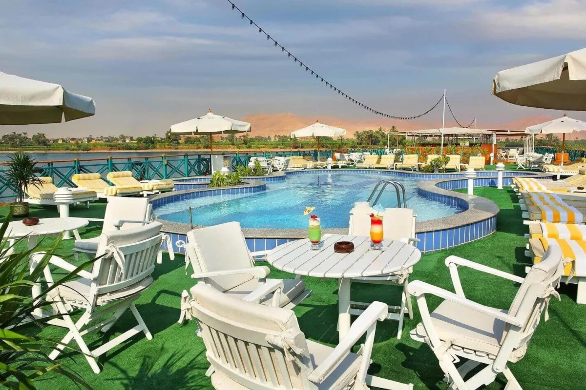 White timber chairs and tables around the pool on the Sonesta Moon Goddess Nile cruise sun deck, dunes on the far bank