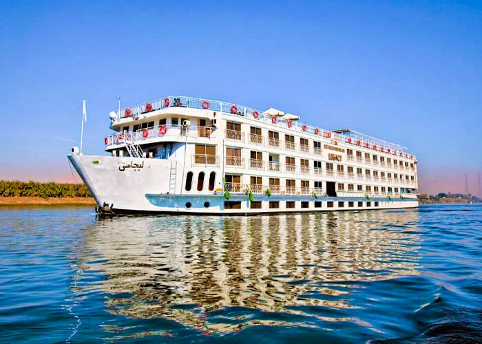 The Steigenberger Legacy Nile cruise ship on the Nile, its name in Arabic on the bow and reeds along the far bank