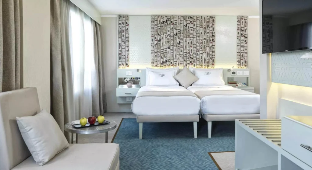 A twin-bed cabin on the Tulip Nile cruise with white linen, a mosaic feature wall, a sofa by the window and a wall-mounted television